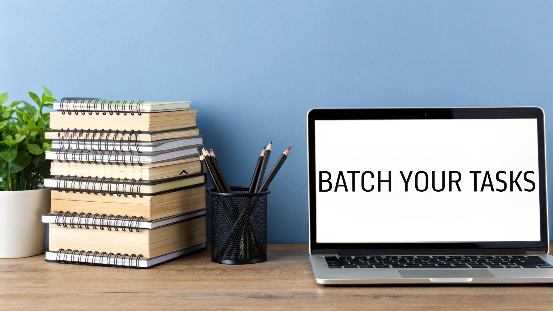 A tidy workspace with a stack of notebooks, pencils, a plant, and a laptop showing 'BATCH YOUR TASKS'.